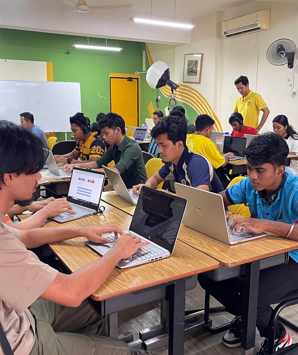 A digital skills class in progress: a room of young adults at shared desks, each working on a laptop with headphones on, while a trainer moves between them.
